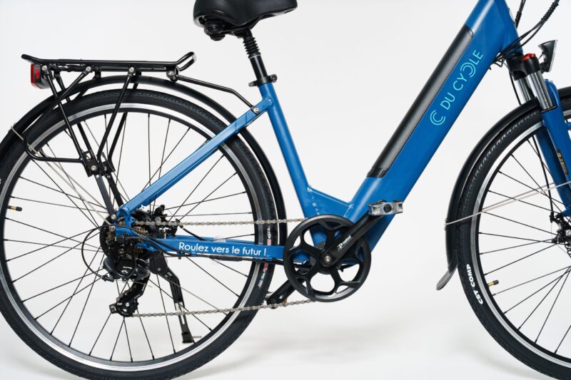 Vélo électrique Ariane bleu mat, vue de la face droite, zoom sur dérailleur arrière, pédales, porte bagage, selle suspendue, béquille, cadre avec batterie intégrée et enjambement bas